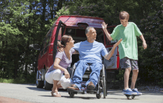 man on wheelchair outdoors with wife kneeling beside him and son on skateboard giving a high five